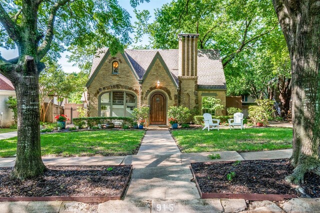 tudor home with a front lawn, a shingled roof, a chimney, and brick siding