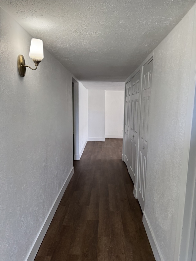 hall with dark wood-type flooring, a textured wall, baseboards, and a textured ceiling