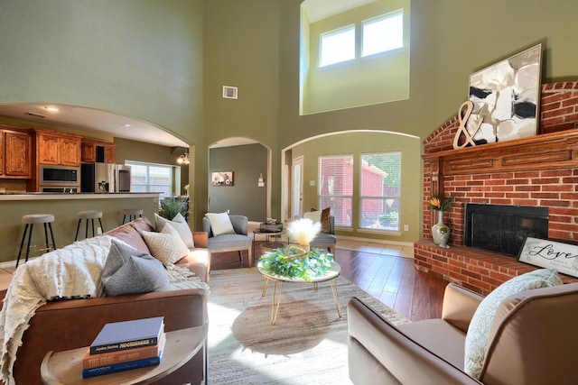 living room featuring wood-type flooring, a brick fireplace, visible vents, a towering ceiling, and arched walkways