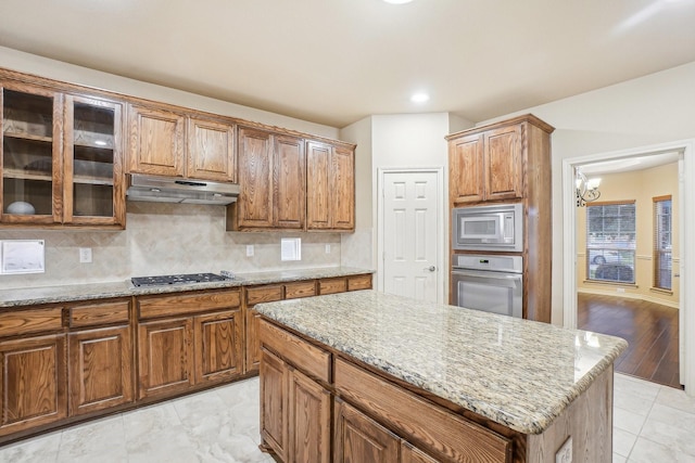 kitchen featuring light stone countertops, stainless steel appliances, under cabinet range hood, and brown cabinetry