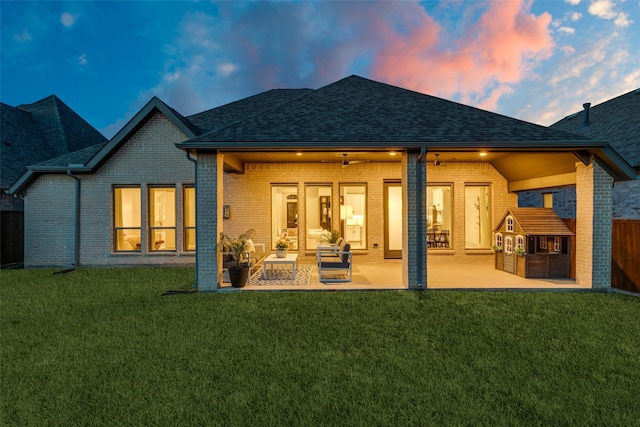 back of house at dusk featuring a lawn, brick siding, fence, and roof with shingles