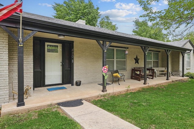 entrance to property featuring a chimney, roof with shingles, a yard, a porch, and brick siding