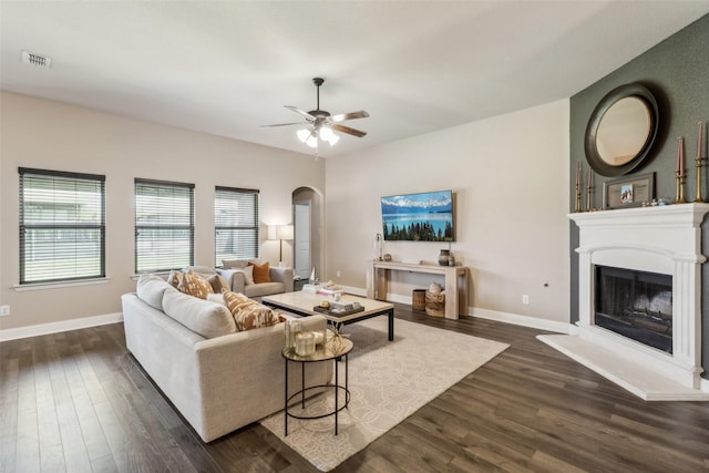 living area with arched walkways, a ceiling fan, dark wood-style flooring, and a fireplace with raised hearth