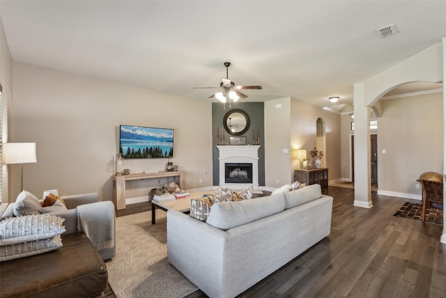 living area with dark wood-style flooring, a fireplace, ceiling fan, baseboards, and arched walkways