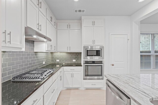7416 San Jacinto Trl, Mckinney, TX 75071 - kitchen with stainless steel appliances, under cabinet range hood, dark stone counters, white cabinetry, and decorative backsplash