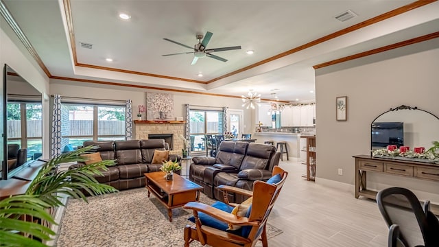 living room featuring a tray ceiling, visible vents, a stone fireplace, ornamental molding, and ceiling fan with notable chandelier