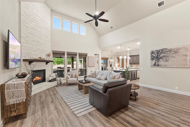 living room with visible vents, wood finished floors, high vaulted ceiling, a stone fireplace, and ceiling fan with notable chandelier