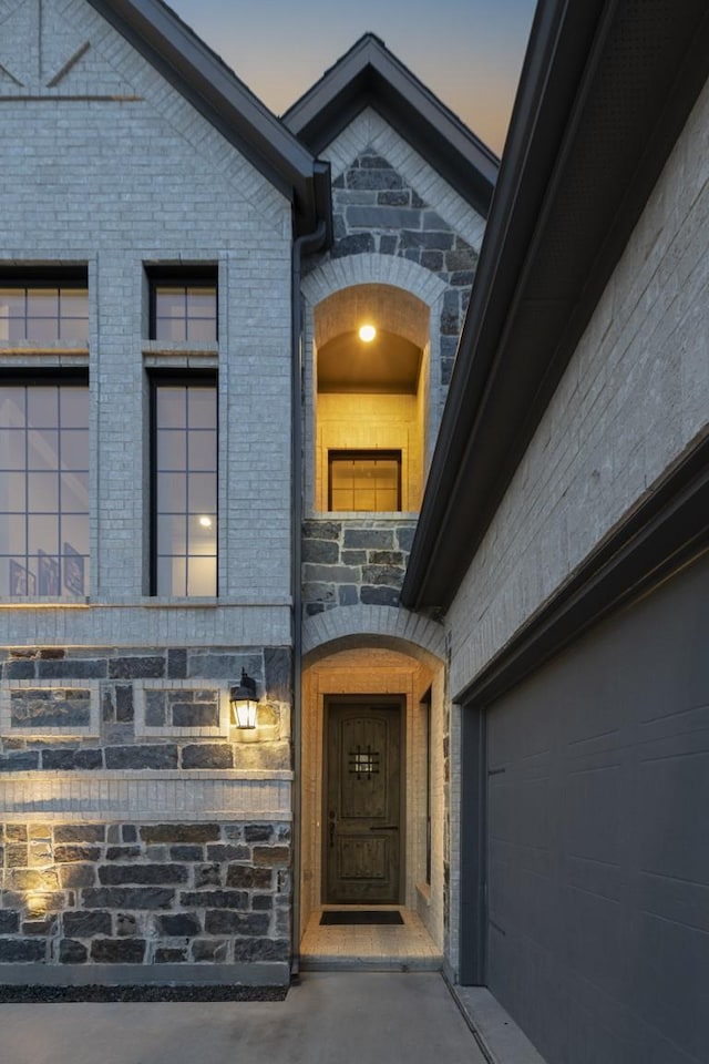 exterior entry at dusk featuring a garage, stone siding, and brick siding