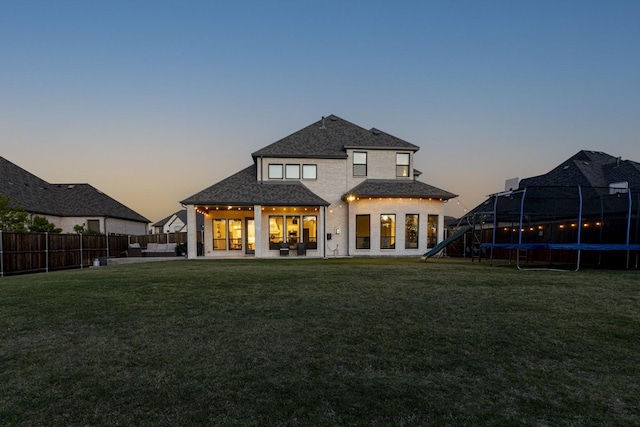 back of property at dusk featuring a patio, a fenced backyard, roof with shingles, a lawn, and a trampoline