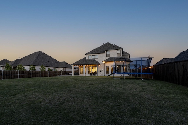 back of house at dusk with a lawn, a fenced backyard, and a trampoline