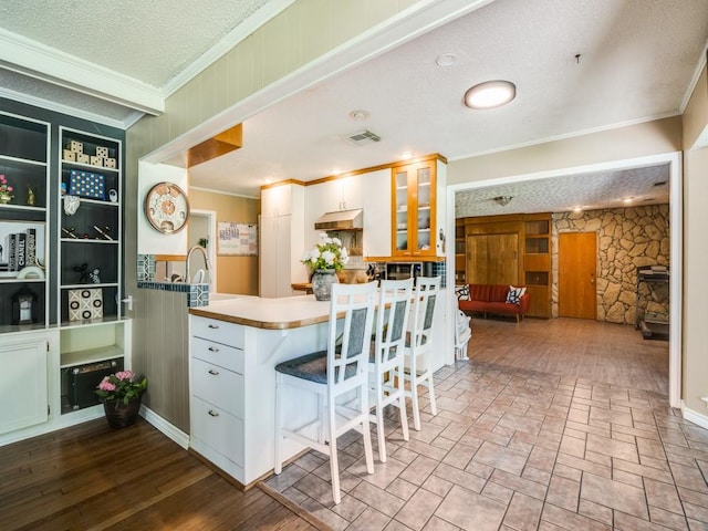 kitchen featuring under cabinet range hood, glass insert cabinets, crown molding, visible vents, and a peninsula