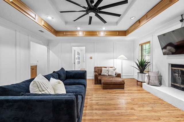 living room featuring ceiling fan, light wood-style floors, a tray ceiling, a glass covered fireplace, and a decorative wall