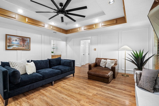 living room featuring light wood finished floors, recessed lighting, ceiling fan, a raised ceiling, and a decorative wall