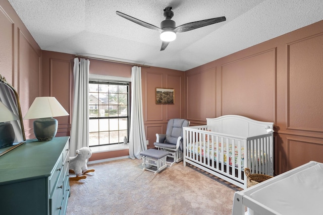 bedroom featuring a textured ceiling, light colored carpet, ceiling fan, a crib, and a decorative wall