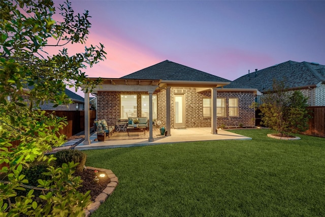 back of house at dusk with a fenced backyard, brick siding, a patio, roof with shingles, and a lawn