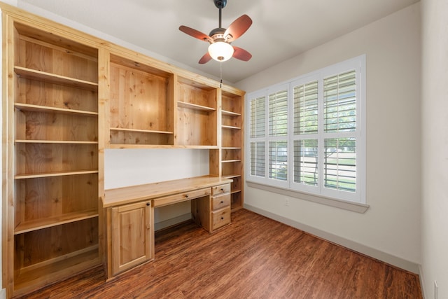 unfurnished office with built in desk, baseboards, a ceiling fan, and dark wood-type flooring