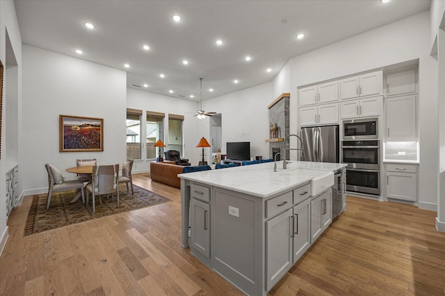 kitchen with light stone counters, stainless steel appliances, light wood-type flooring, a kitchen island with sink, and gray cabinets