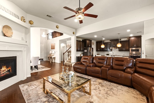 living room with a tiled fireplace, a ceiling fan, visible vents, arched walkways, and wood finished floors