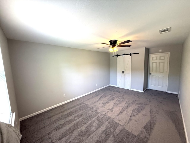 unfurnished bedroom with dark carpet, a barn door, visible vents, baseboards, and a ceiling fan