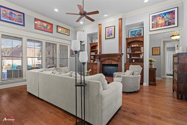 living room with a ceiling fan, crown molding, built in features, dark wood-style floors, and a glass covered fireplace