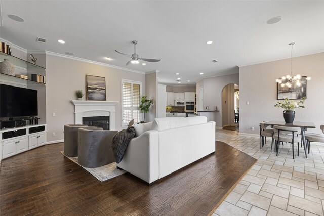 living area with crown molding, arched walkways, a glass covered fireplace, ceiling fan with notable chandelier, and recessed lighting