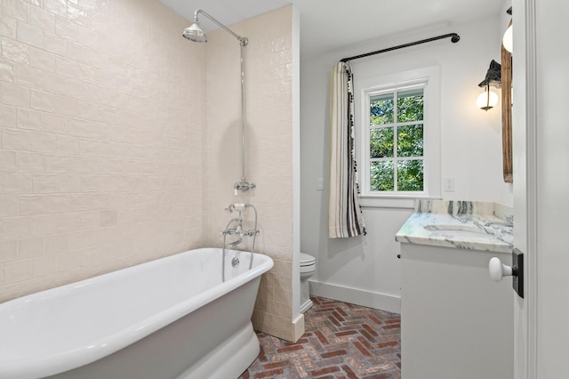 bathroom featuring vanity, toilet, a freestanding bath, baseboards, and brick floor