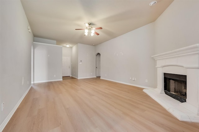 unfurnished living room featuring arched walkways, light wood-style floors, a fireplace with raised hearth, baseboards, and a ceiling fan