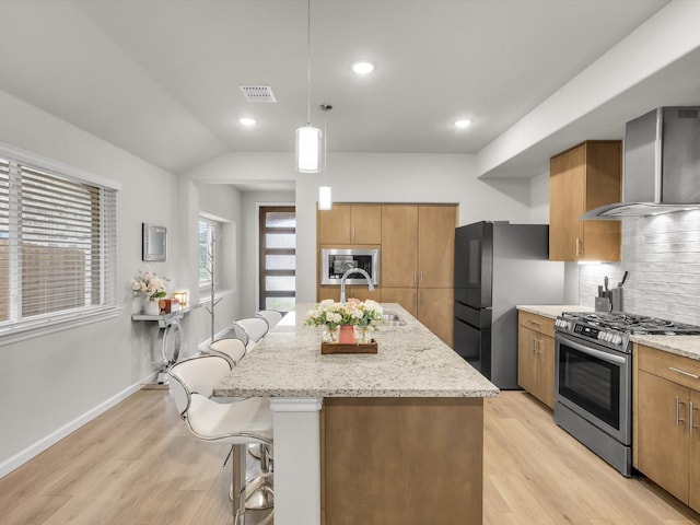 kitchen with light wood finished floors, brown cabinetry, wall chimney exhaust hood, a breakfast bar, and appliances with stainless steel finishes