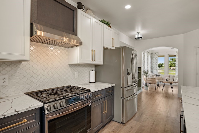 kitchen featuring appliances with stainless steel finishes, light stone counters, wall chimney range hood, and arched walkways