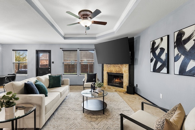living room with a warm lit fireplace, light carpet, baseboards, a raised ceiling, and a ceiling fan
