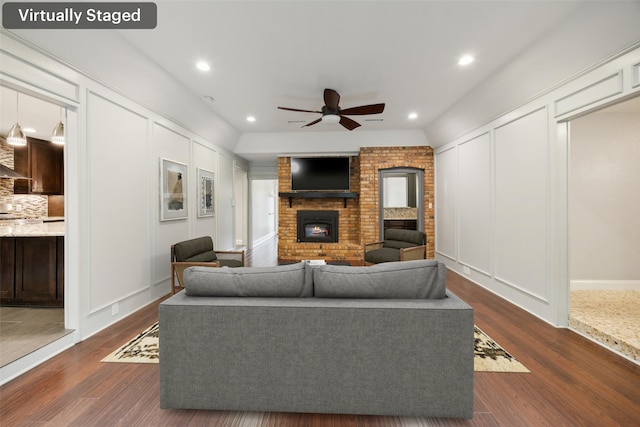 living area featuring a brick fireplace, ceiling fan, dark wood-type flooring, and a decorative wall