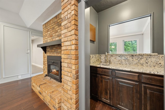 kitchen with a brick fireplace, dark wood finished floors, dark brown cabinets, a sink, and light stone counters