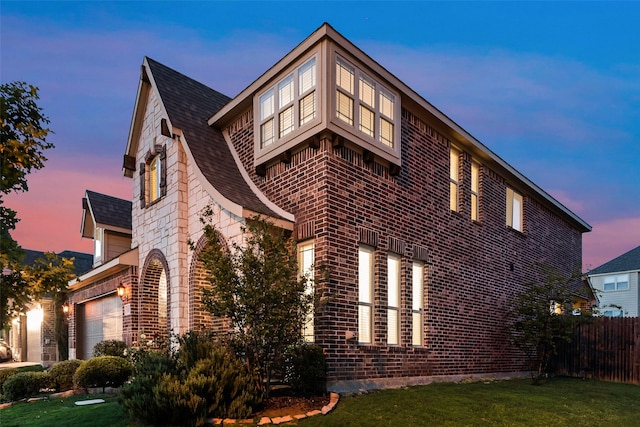 property exterior at dusk with a shingled roof, fence, a yard, brick siding, and a garage