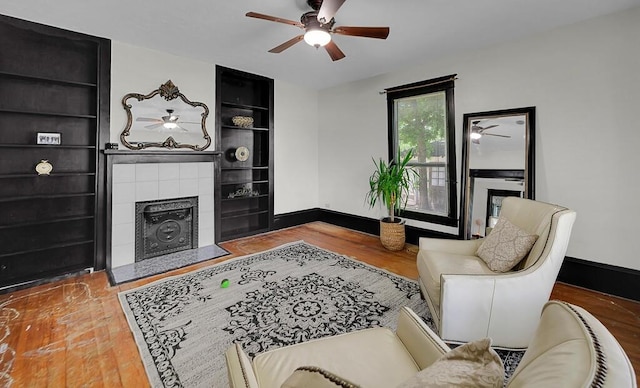 living area featuring a tile fireplace, built in features, a ceiling fan, baseboards, and wood finished floors