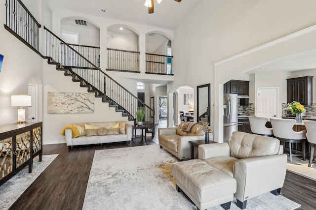 living area with arched walkways, ceiling fan, dark wood-style flooring, stairway, and a towering ceiling