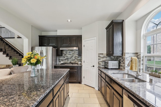 kitchen with dark stone countertops, decorative backsplash, a sink, stainless steel appliances, and under cabinet range hood