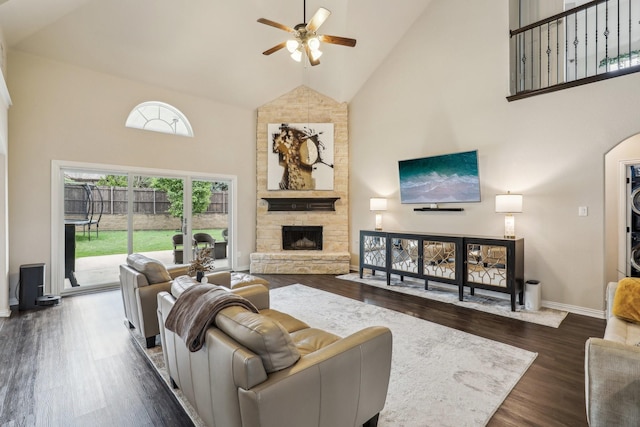 living room with dark wood-style floors, high vaulted ceiling, a ceiling fan, a stone fireplace, and baseboards