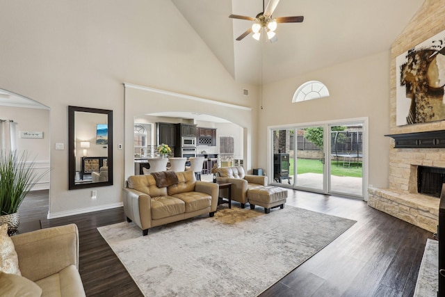 living area featuring dark wood-style flooring, ceiling fan, high vaulted ceiling, a stone fireplace, and arched walkways