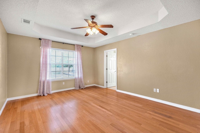 empty room with a textured ceiling, light wood-style floors, a tray ceiling, baseboards, and a ceiling fan