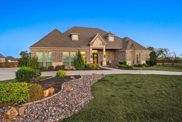 french country home with a front lawn, fence, roof with shingles, and brick siding