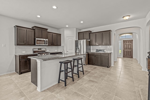 kitchen featuring appliances with stainless steel finishes, stone counters, backsplash, arched walkways, and dark brown cabinets