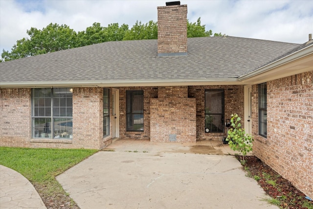 property entrance with brick siding, a shingled roof, a patio area, and a chimney
