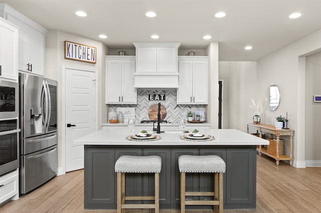 kitchen with a kitchen island with sink, stainless steel appliances, light wood-style floors, backsplash, and white cabinets