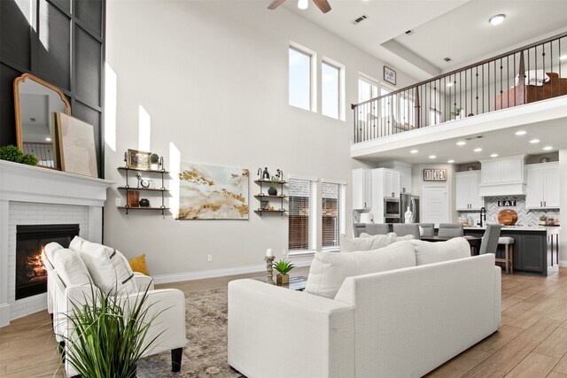 living area with ceiling fan, a brick fireplace, baseboards, light wood-type flooring, and a towering ceiling