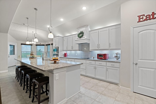kitchen featuring an island with sink, custom range hood, appliances with stainless steel finishes, backsplash, and light tile patterned flooring
