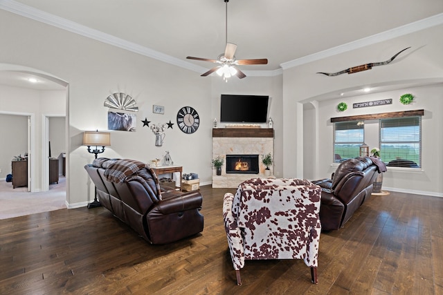 living room featuring arched walkways, a stone fireplace, wood-type flooring, a ceiling fan, and crown molding