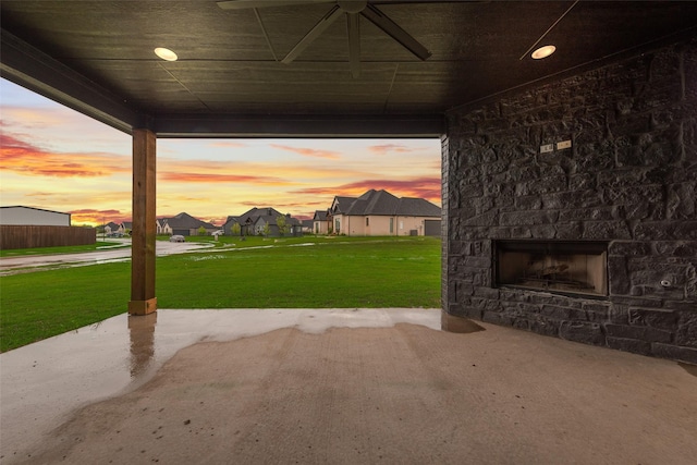 patio terrace at dusk with a yard, an outdoor stone fireplace, a residential view, and a ceiling fan