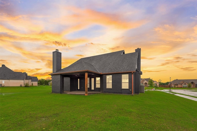 back of house at dusk featuring brick siding, a chimney, cooling unit, a lawn, and a patio
