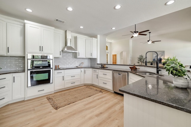 kitchen with light wood-style flooring, decorative backsplash, appliances with stainless steel finishes, a sink, and wall chimney exhaust hood