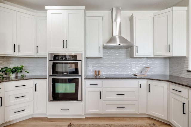 kitchen featuring double oven, dark stone counters, white cabinetry, and wall chimney exhaust hood